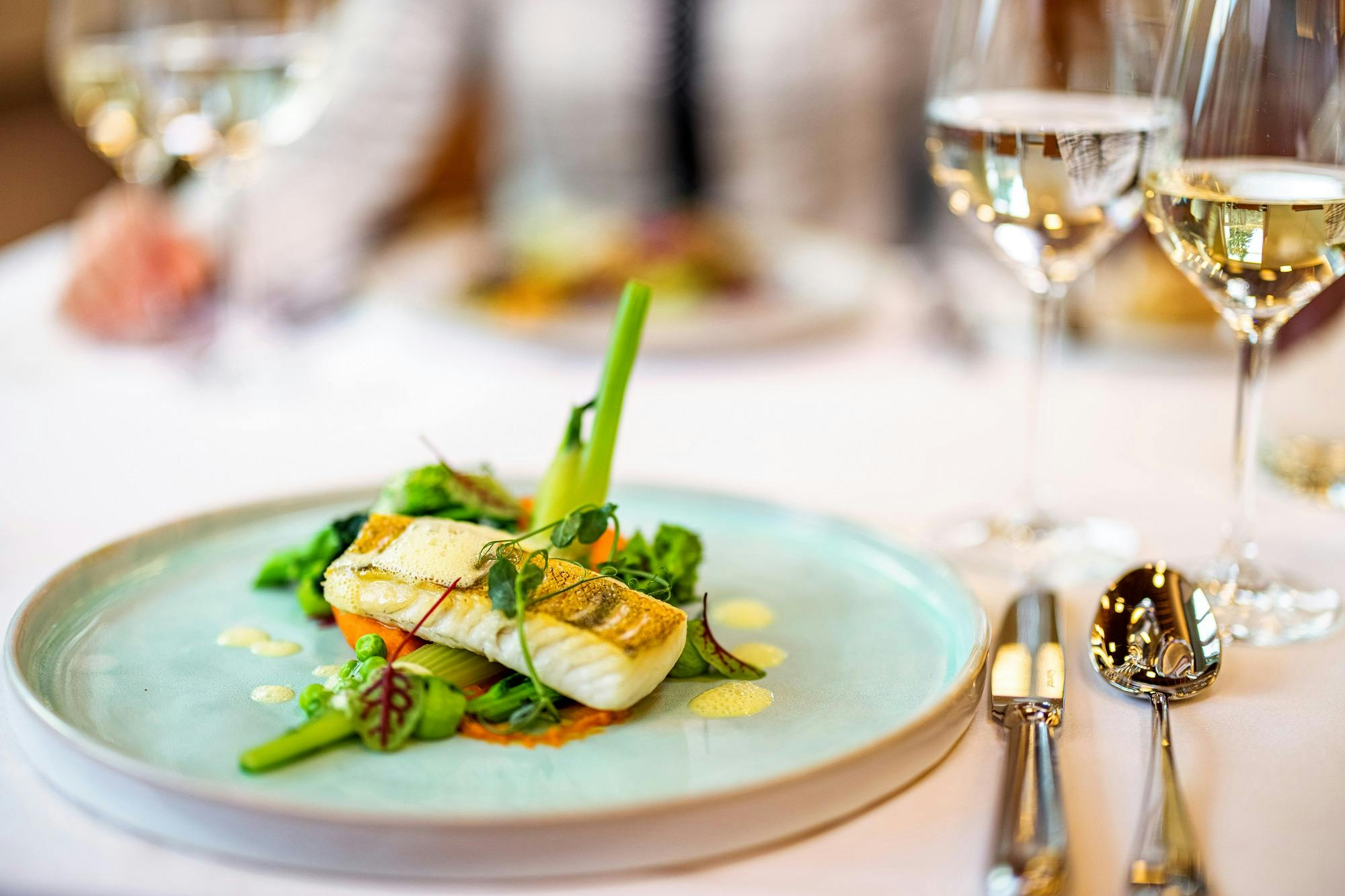 Elegantes Gourmet-Essen mit Fischfilet und frischem Gemüse auf einem Teller, begleitet von einem Glas Weißwein.