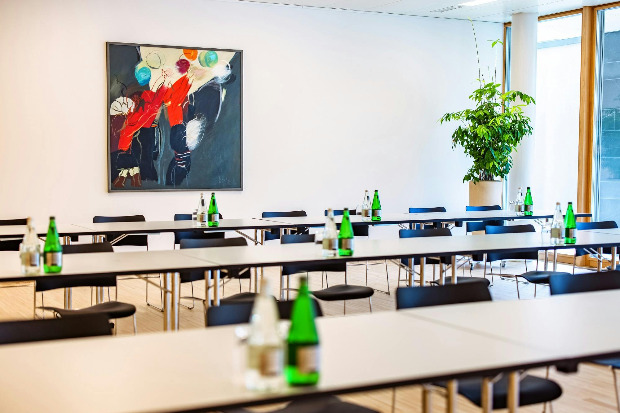 Moderner Besprechungsraum mit Tischen, Stühlen und grünen Wasserflaschen, dekoriert mit einem großen Gemälde und einer Zimmerpflanze.